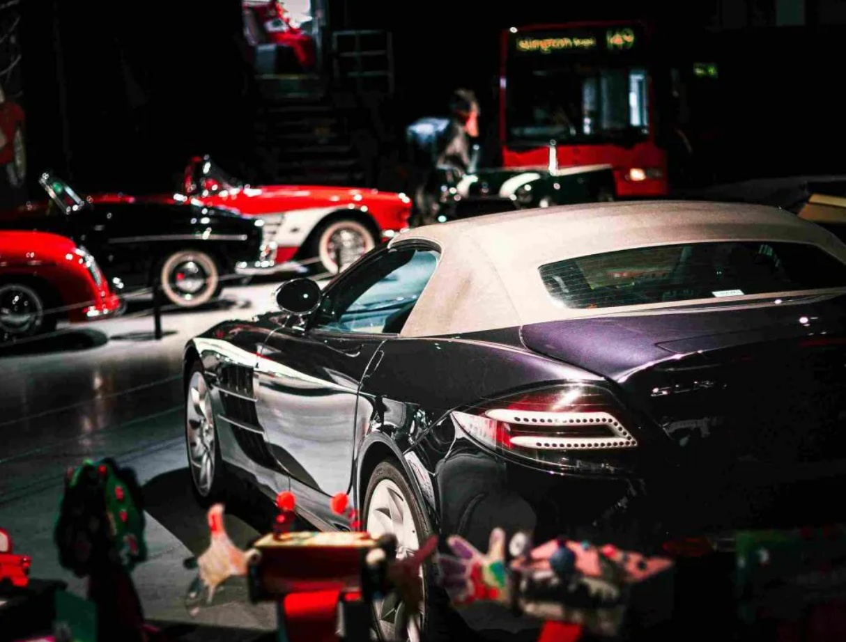 A selection of classic and modern cars on indoor display, with a black Mercedes-Benz sports car in the foreground and several vintage cars and a red double-decker bus in the background